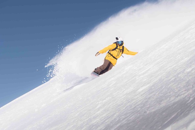 Person in a yellow jacket snowboarding down a snowy slope, with a clear blue sky in the background